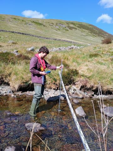 A man stands in a shallow river with a metal stick to monitor the river flow