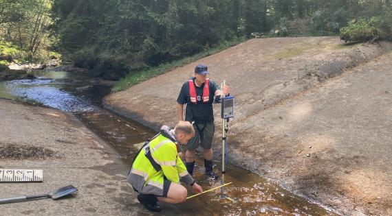 Two men stand in a shallow river with a metal stick to monitor the river flow