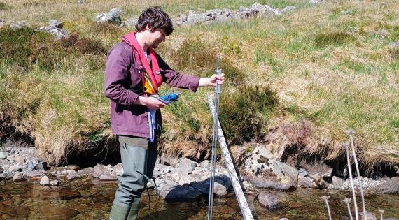 A man stands in a shallow river with a metal stick to monitor the river flow