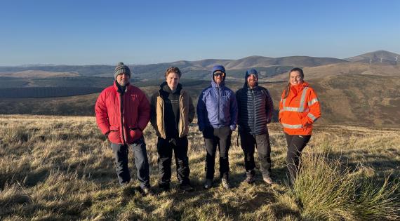 Five colleagues stand in the Tweed catchment, with a hilly backdrop behind them