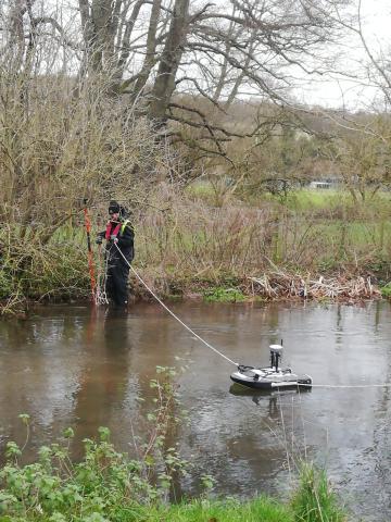 A hydrologist stands in a shallow river using a remote control boat to measure the speed of the water