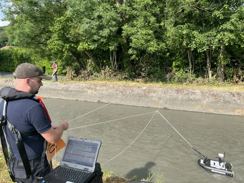 A man stands beside a river, with a rope suspending a hydroboard on the surface