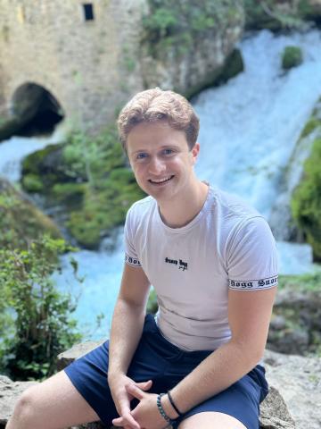 Image shows PhD candidate, Alex Braggins, smiling and sitting on a rock next to a waterfall