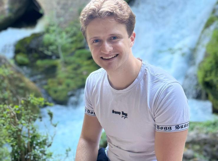 Image shows PhD candidate, Alex Braggins, smiling and sitting on a rock next to a waterfall
